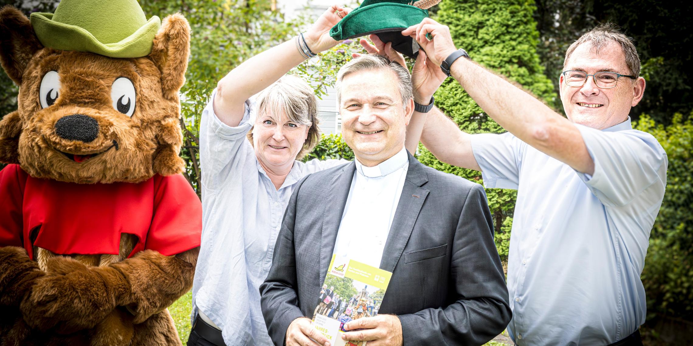 Gruppenfoto mit Maskottchen Robbi, Andrea Elsmann, Stadtdechant Hofmann und Jean-Pierre Schneider. Die beiden Geschäftsführer von Diakonie und Caritas setzen dem neuen Paten Dr. Hofmann symbolisch eine ROBIN GOOD Mütze auf.