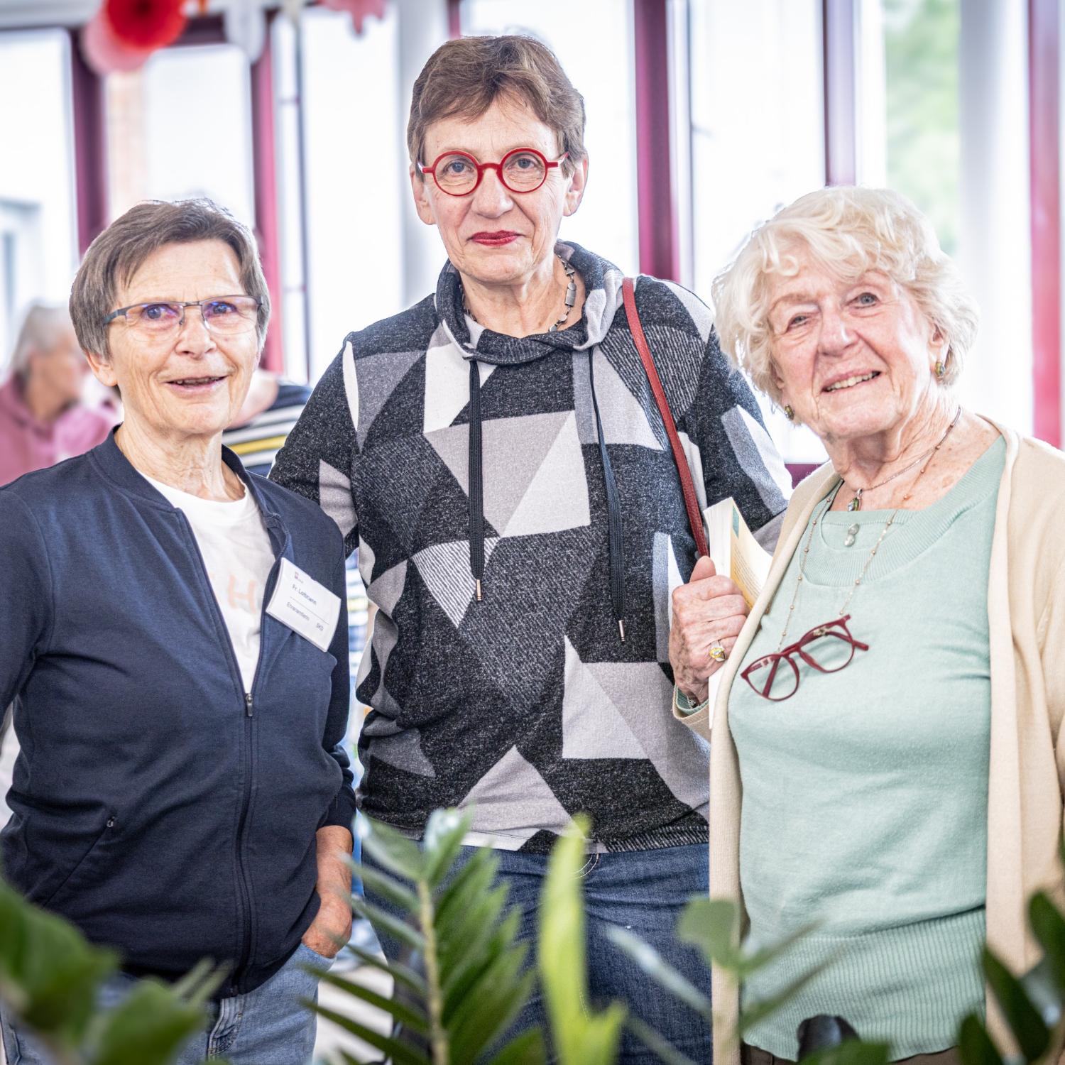 Drei ältere Frauen stehen lächelnd beisammen in einem hellen Aufenthaltsraum des Altenheims Herz Jesu Kloster. Sie wirken freundlich und engagiert – als ehrenamtliche Helferinnen – und tragen teils Namensschilder oder Bücher, umgeben von weiteren Senior\*innen im Hintergrund.