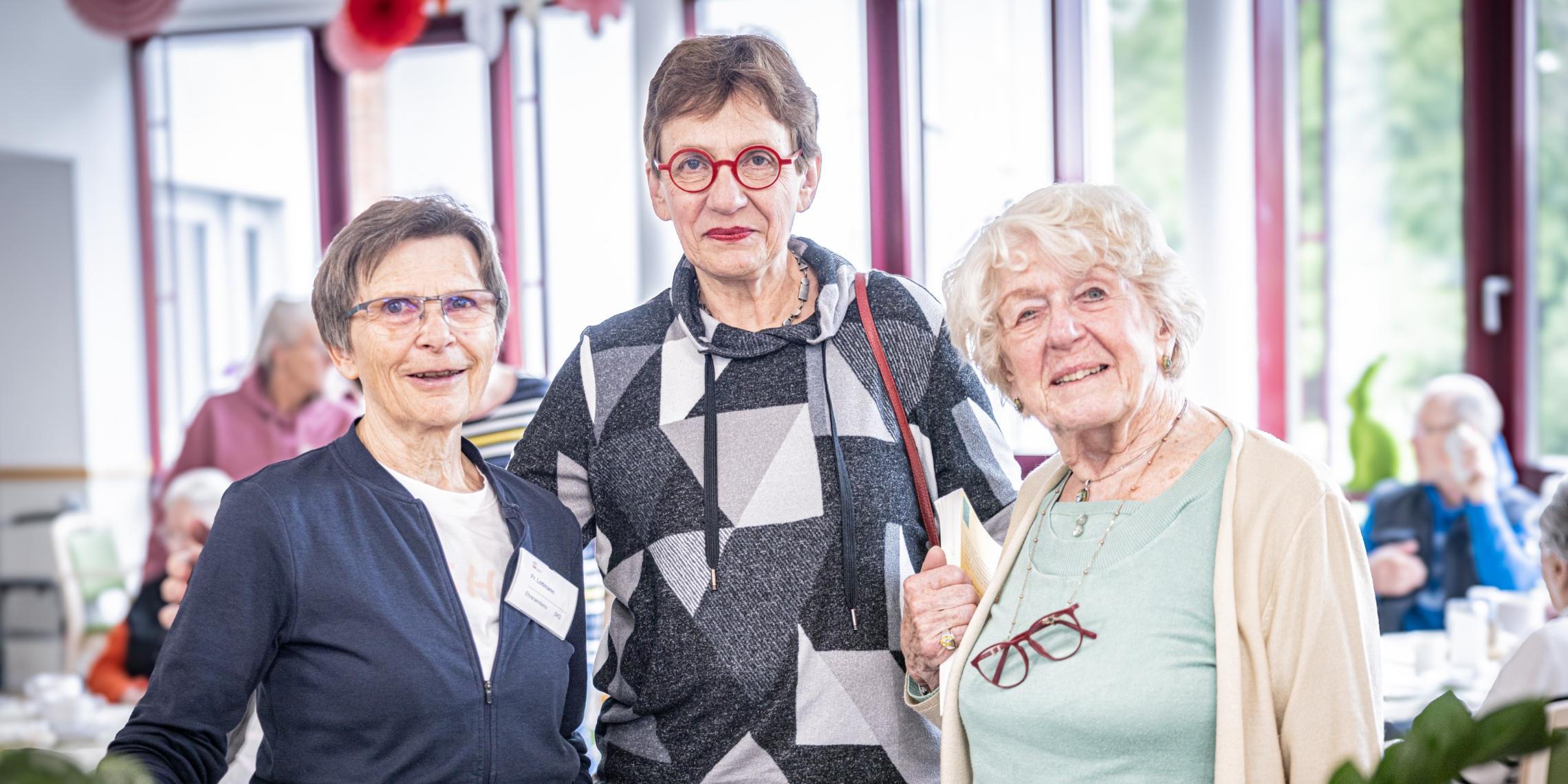 Drei ältere Frauen stehen lächelnd beisammen in einem hellen Aufenthaltsraum des Altenheims Herz Jesu Kloster. Sie wirken freundlich und engagiert – als ehrenamtliche Helferinnen – und tragen teils Namensschilder oder Bücher, umgeben von weiteren Senior\*innen im Hintergrund.