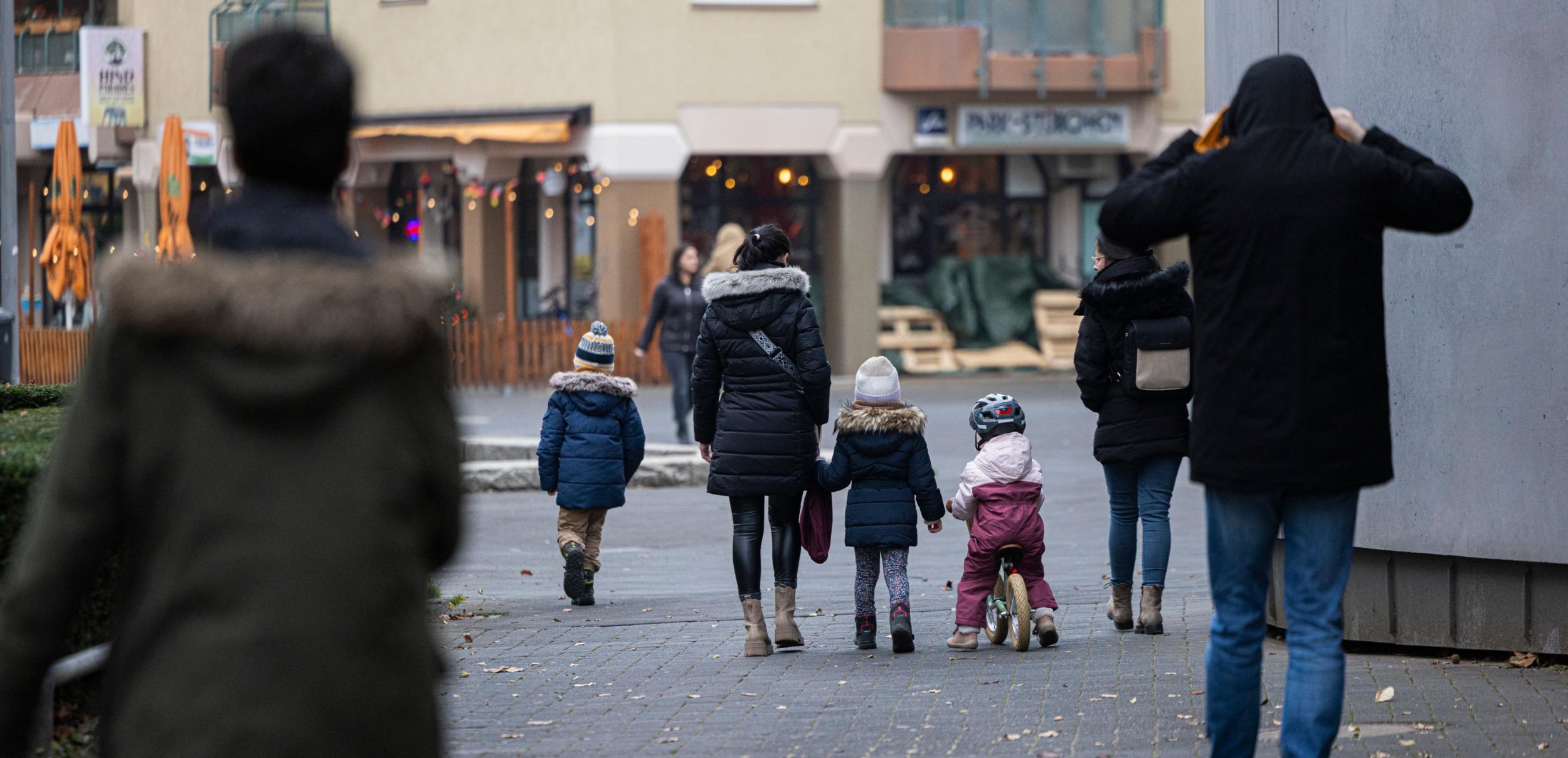 Eine Straßenszene in einer Wohngegend an einem kühlen Tag. Mehrere Erwachsene und Kinder in Winterkleidung gehen auf einem gepflasterten Weg, einige halten sich an den Händen. Ein Kind fährt auf einem kleinen Laufrad, während im Hintergrund beleuchtete Schaufenster und eine Außengastronomie mit geschlossenen Sonnenschirmen zu sehen sind.