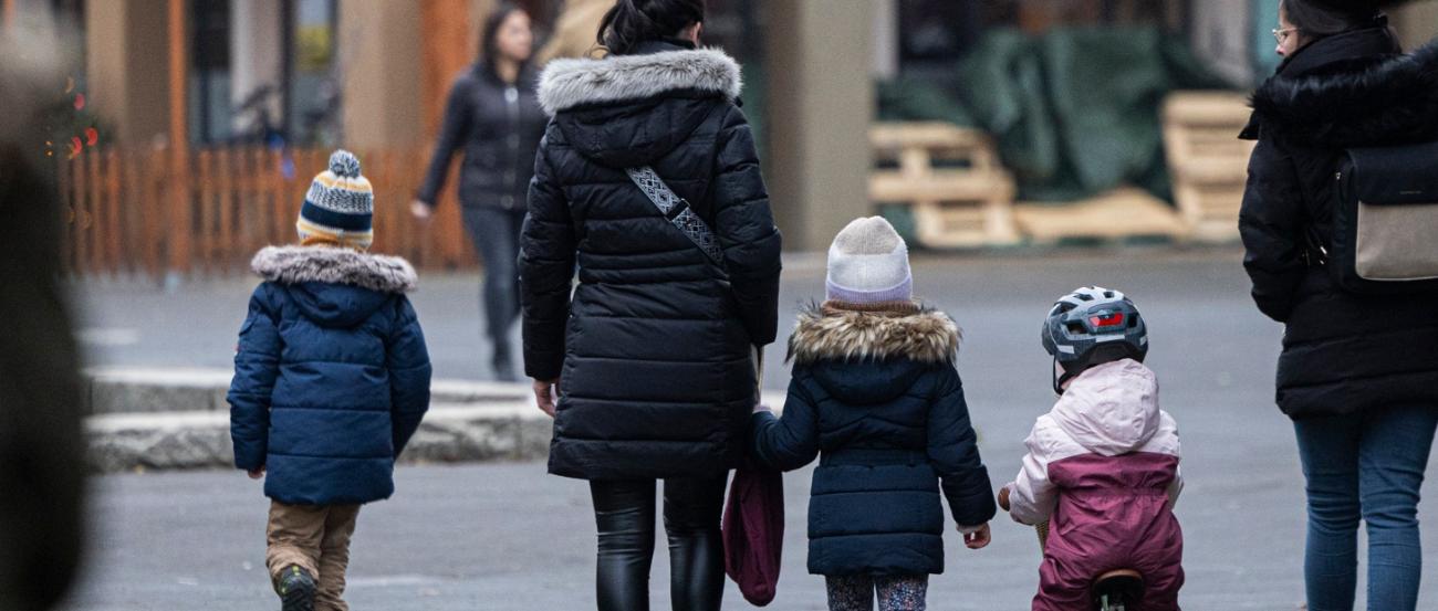 Eine Straßenszene in einer Wohngegend an einem kühlen Tag. Mehrere Erwachsene und Kinder in Winterkleidung gehen auf einem gepflasterten Weg, einige halten sich an den Händen. Ein Kind fährt auf einem kleinen Laufrad, während im Hintergrund beleuchtete Schaufenster und eine Außengastronomie mit geschlossenen Sonnenschirmen zu sehen sind.