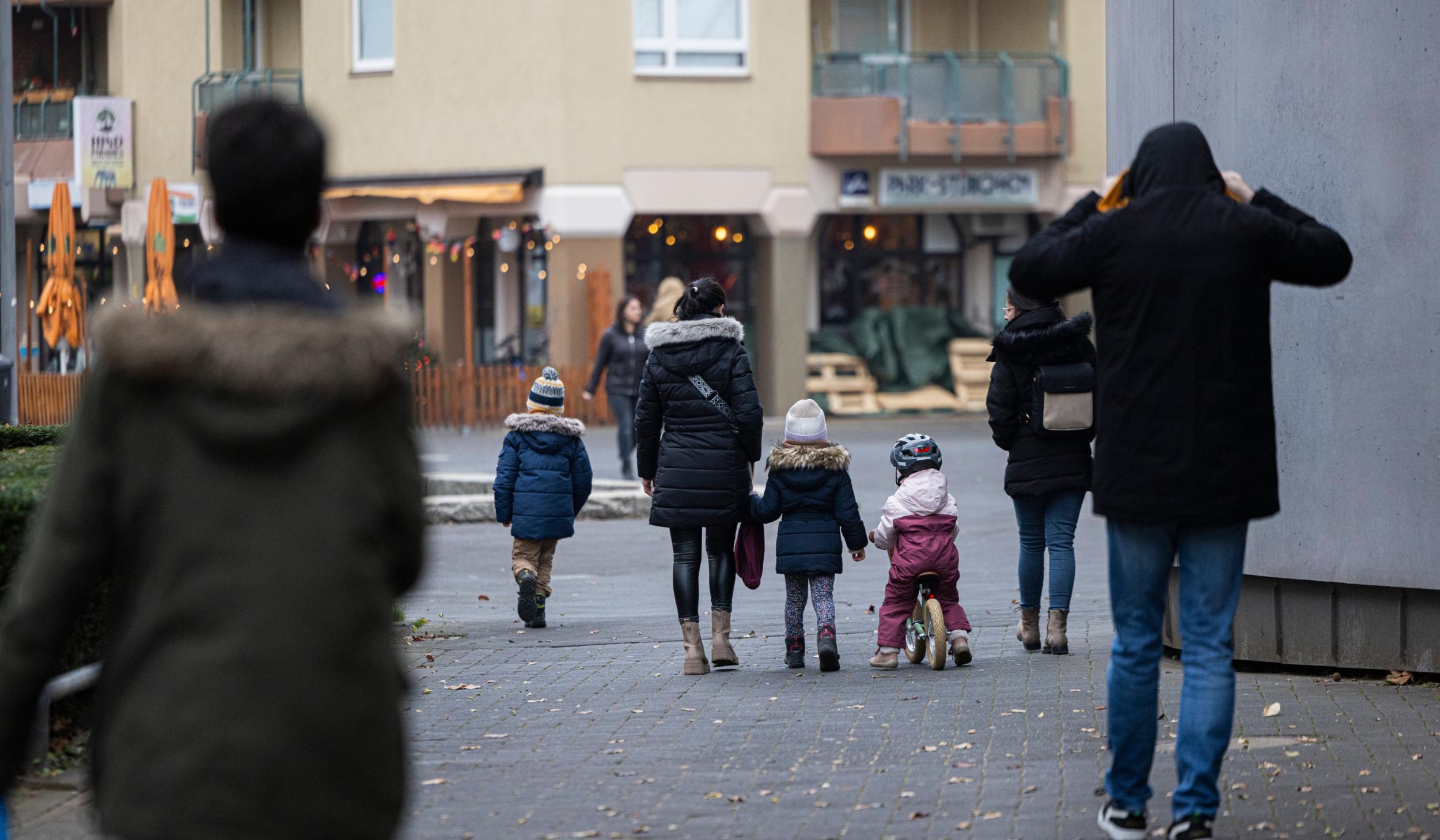 Eine Straßenszene in einer Wohngegend an einem kühlen Tag. Mehrere Erwachsene und Kinder in Winterkleidung gehen auf einem gepflasterten Weg, einige halten sich an den Händen. Ein Kind fährt auf einem kleinen Laufrad, während im Hintergrund beleuchtete Schaufenster und eine Außengastronomie mit geschlossenen Sonnenschirmen zu sehen sind.