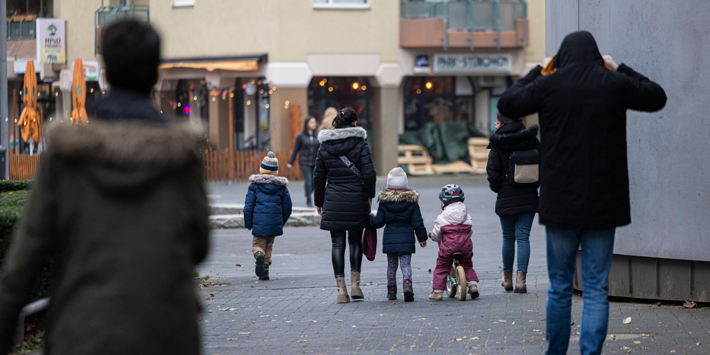 Eine Straßenszene in einer Wohngegend an einem kühlen Tag. Mehrere Erwachsene und Kinder in Winterkleidung gehen auf einem gepflasterten Weg, einige halten sich an den Händen. Ein Kind fährt auf einem kleinen Laufrad, während im Hintergrund beleuchtete Schaufenster und eine Außengastronomie mit geschlossenen Sonnenschirmen zu sehen sind.