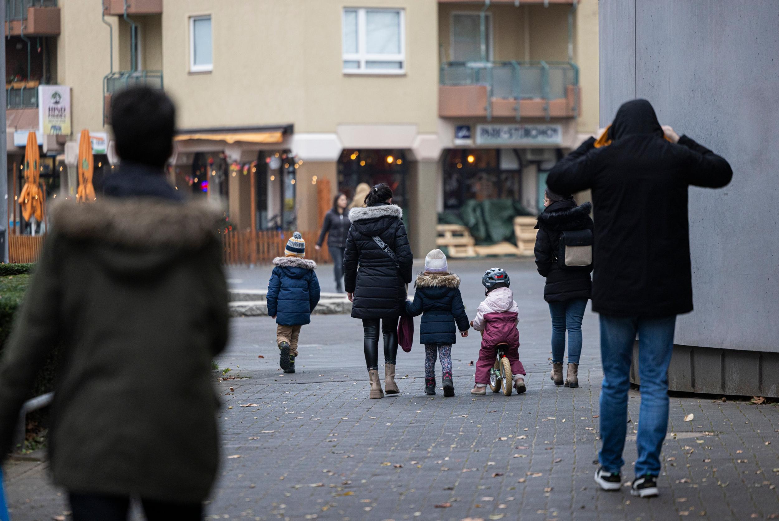 Eine Straßenszene in einer Wohngegend an einem kühlen Tag. Mehrere Erwachsene und Kinder in Winterkleidung gehen auf einem gepflasterten Weg, einige halten sich an den Händen. Ein Kind fährt auf einem kleinen Laufrad, während im Hintergrund beleuchtete Schaufenster und eine Außengastronomie mit geschlossenen Sonnenschirmen zu sehen sind.